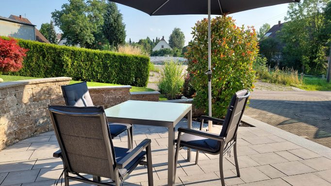 Ein Tisch mit Stühlen unter einem Sonnenschirm in einem Garten mit grüner Umgebung.