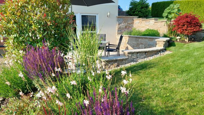 Blühender Garten mit bunten Pflanzen, Sitzbereich und einem Haus im Hintergrund.