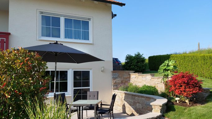 Haus mit Terrasse, Sonnenschirm und blühenden Sträuchern im Garten.